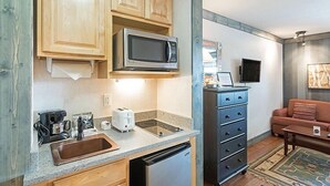 A compact kitchen with wooden cabinets, a microwave, a toaster, a sink, and a dishwasher. There's a black cabinet with drawers, a brown couch, and a rug in the living area. - Mountainside Inn 116 1 Bedroom Hotel Room by Alpine Lodging Telluride (Telluride)