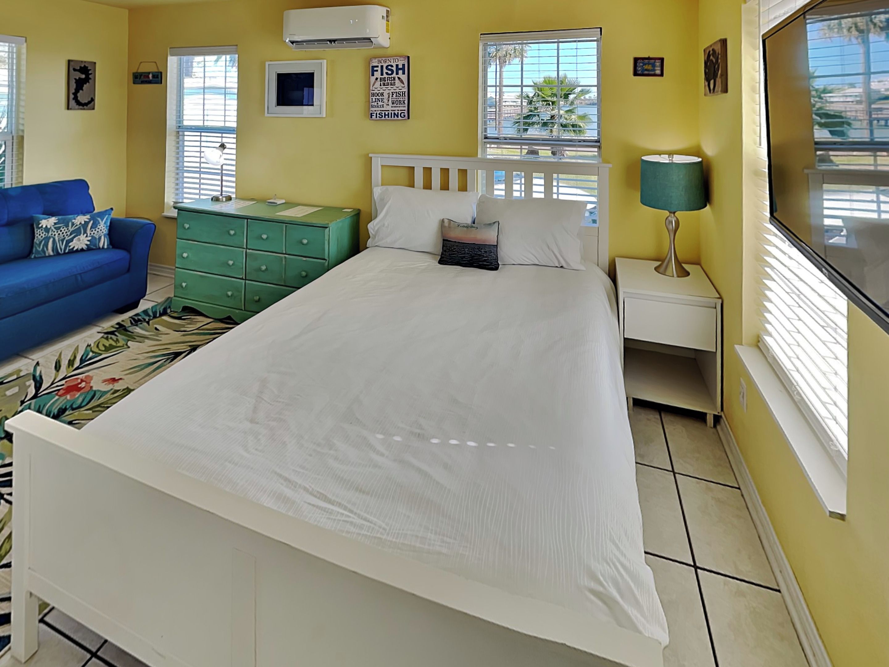 A bedroom with a bed, a nightstand, a lamp, a dresser, a blue sofa, and a window with blinds.