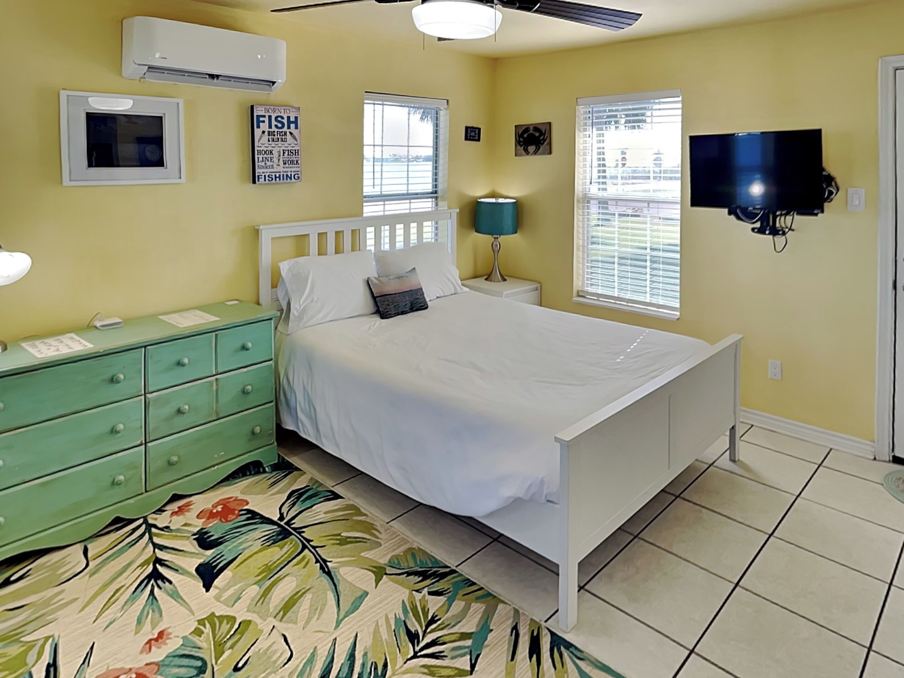 A bedroom with a bed, a green dresser, a wall-mounted TV, and a window with blinds.