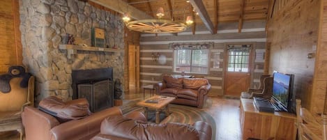 Un interior de cabaña acogedor con pared de piedra, sofás de cuero, chimenea y televisor.
