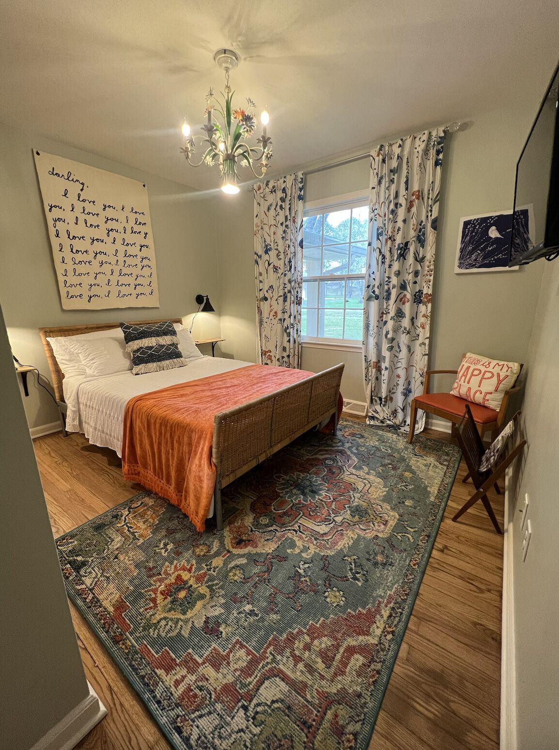 Une chambre à coucher avec un lit, une table de chevet, une chaise, une fenêtre avec des rideaux, un lustre et un mur sur lequel est écrit un message.
