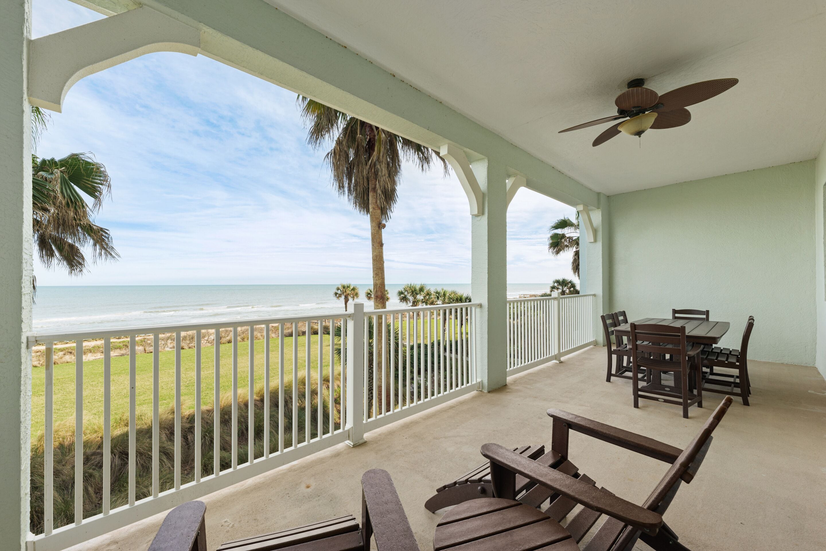 A covered outdoor area with a view of the ocean, a table and chairs, and a ceiling fan.