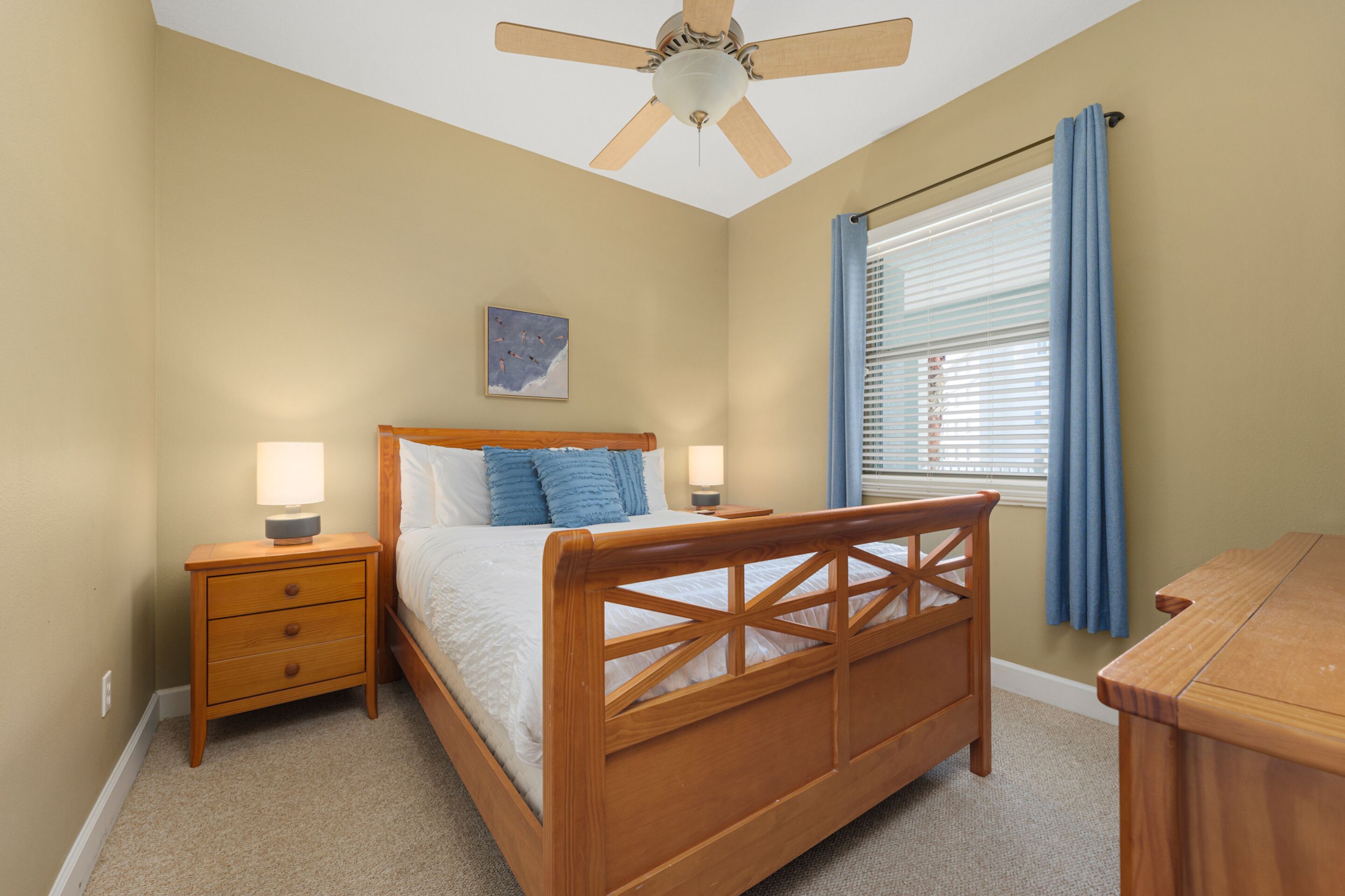 Une chambre à coucher avec un lit en bois, une table de chevet avec une lampe, un ventilateur de plafond et une fenêtre avec des rideaux.