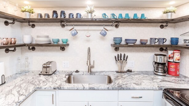A kitchen with a marble countertop, stainless steel sink, and various kitchen utensils. There are shelves above the countertop holding cups and bowls.