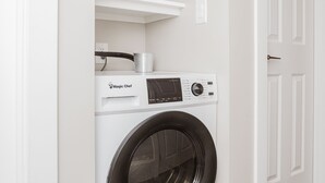 A compact laundry room with a front-loading washing machine, a shelf with folded towels, and a white door.
