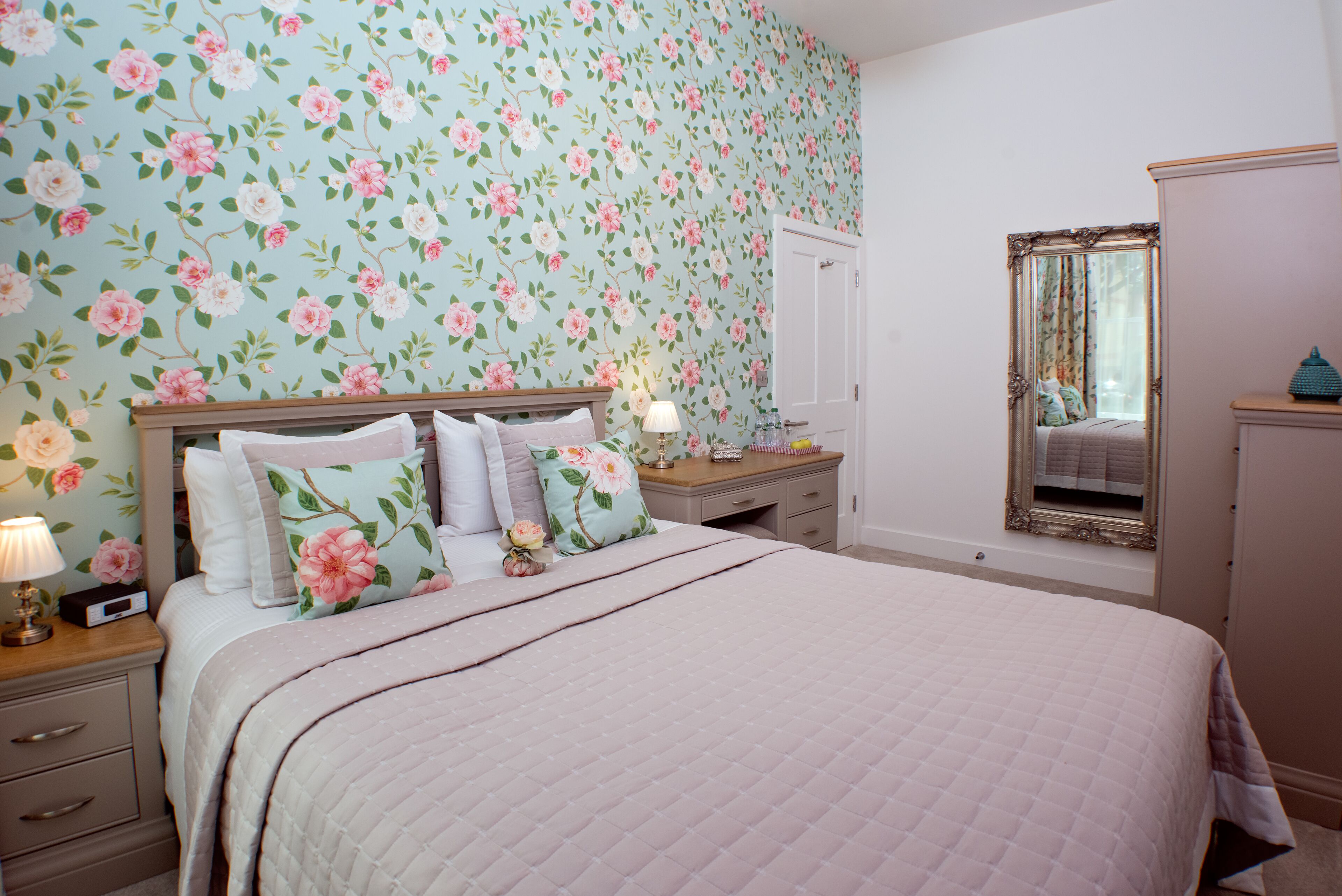 A bedroom with floral wallpaper, a bed with multiple pillows, a nightstand with a lamp, a dresser, and a mirror.