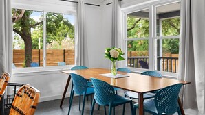 A dining area with a wooden table and blue chairs, large windows with white curtains, and a view of a tree and fence outside.