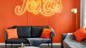 A room with a neon sign reading "Jesse's" on the wall, a black sofa with orange cushions, and a small table with a plant.