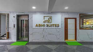 Entrada de un hotel con un muro de mármol, un letrero que dice "ABHI SUITES", un ascensor y una mesa de recepción.