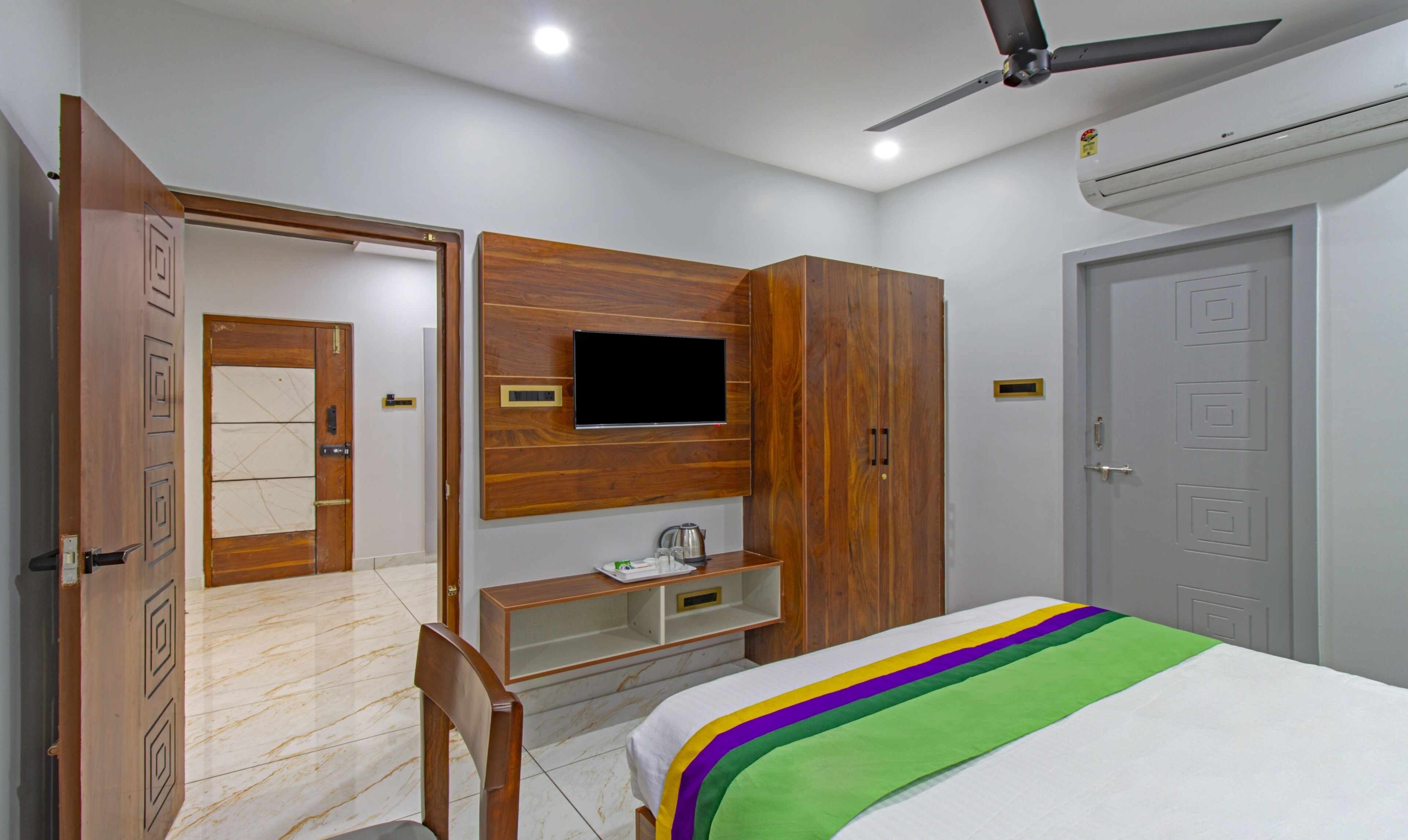 A bedroom with a bed, a TV mounted on the wall, a wardrobe, a desk, and a ceiling fan.