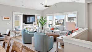A modern living room with a flat-screen TV, two sofas, a dining table, and a ceiling fan. - Osprey Isle: New Build | Private Pool | Rec Room (Corolla)