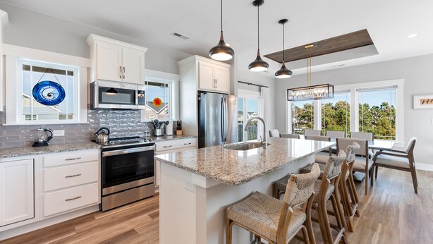 A modern kitchen with white cabinets, stainless steel appliances, a granite countertop, and a large island with bar stools. - Osprey Isle: New Build | Private Pool | Rec Room (Corolla)