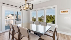 A dining area with a glass-top table, four chairs, and a modern chandelier. - Osprey Isle: New Build | Private Pool | Rec Room (Corolla)