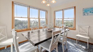 A dining room with a wooden table and white chairs, large windows with blinds, and a chandelier. - SomerVilla: Oceanside | Pool/Hot Tub | Pets OK (Corolla)