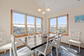 A dining room with a wooden table and white chairs, large windows with blinds, and a chandelier.