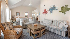 A spacious living room with a high ceiling, wicker furniture, striped sofas, and decorative wall art. - SomerVilla: Oceanside | Pool/Hot Tub | Pets OK (Corolla)