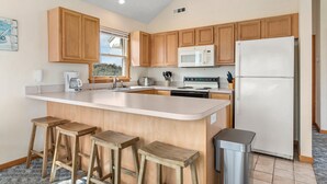 A kitchen with wooden cabinets, a white countertop, a built-in microwave, a refrigerator, and three wooden stools. - SomerVilla: Oceanside | Pool/Hot Tub | Pets OK (Corolla)