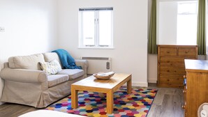 Un salon avec un canapé beige, une table basse en bois, un tapis hexagonal aux couleurs vives, une commode en bois et une fenêtre avec des rideaux.