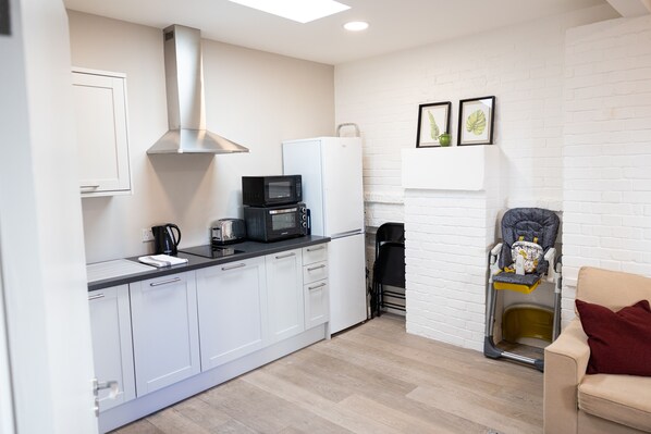 Cocina pequeña con gabinetes blancos, un microondas y un tostador. Hay un cochecito de bebé en la esquina y un sofá color beige con una almohada roja.