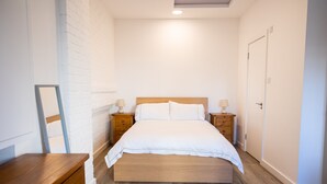 Un dormitorio con cama de madera, ropa de cama blanca, dos mesitas de noche con lámparas, un aparador de madera y una pared de ladrillo blanco.