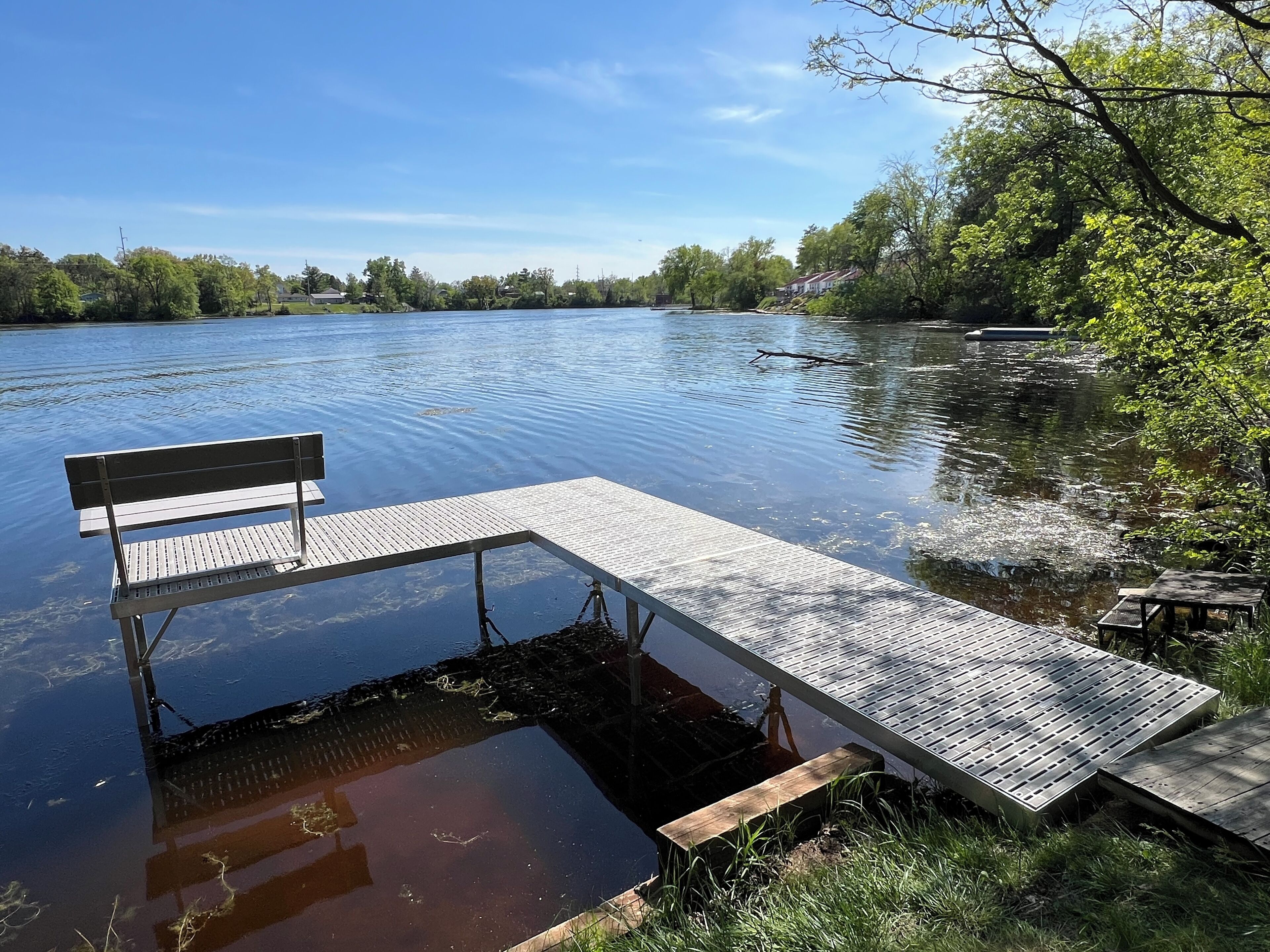 A wooden dock extending into a calm river, surrounded by trees and a clear sky.