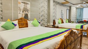 A hotel room with two beds, each with colorful bedding and decorative pillows. There is a wooden headboard, a nightstand, and a small table with a lamp. The room has patterned wallpaper and a large window with curtains. - Treebo Golden Inn, 700 Mtrs From Promenade Beach (Puducherry)