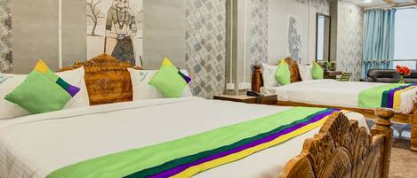Une chambre d’hôtel avec deux lits, chacun agrémenté de draps colorés et de coussins décoratifs. On y trouve une tête de lit en bois, une table de chevet et une petite table avec une lampe. La chambre est décorée de papier peint à motifs et possède une grande fenêtre avec des rideaux.