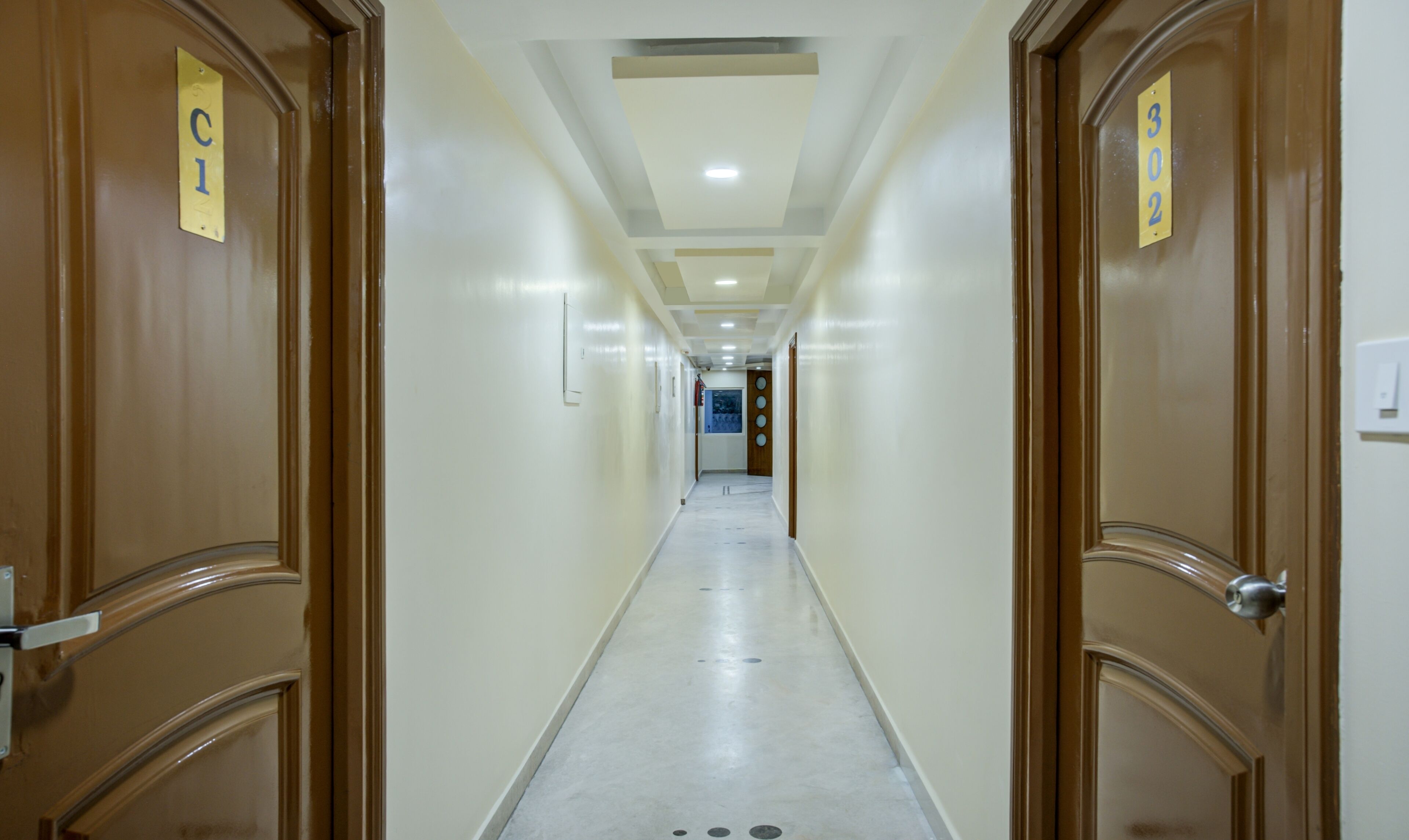 A hotel corridor with wooden doors, a tiled floor, and a ceiling with recessed lighting.