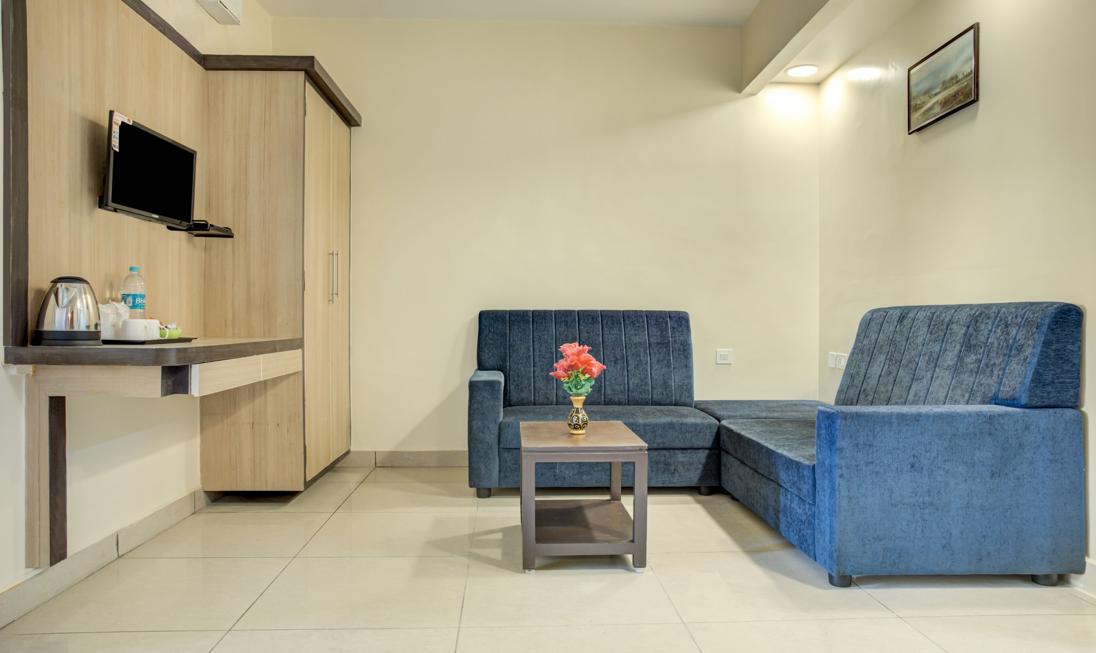 A modern living room with a sectional sofa, a small side table with a vase, a flat-screen TV mounted on a wooden wall, and a kitchenette area with a coffee maker and water bottle.