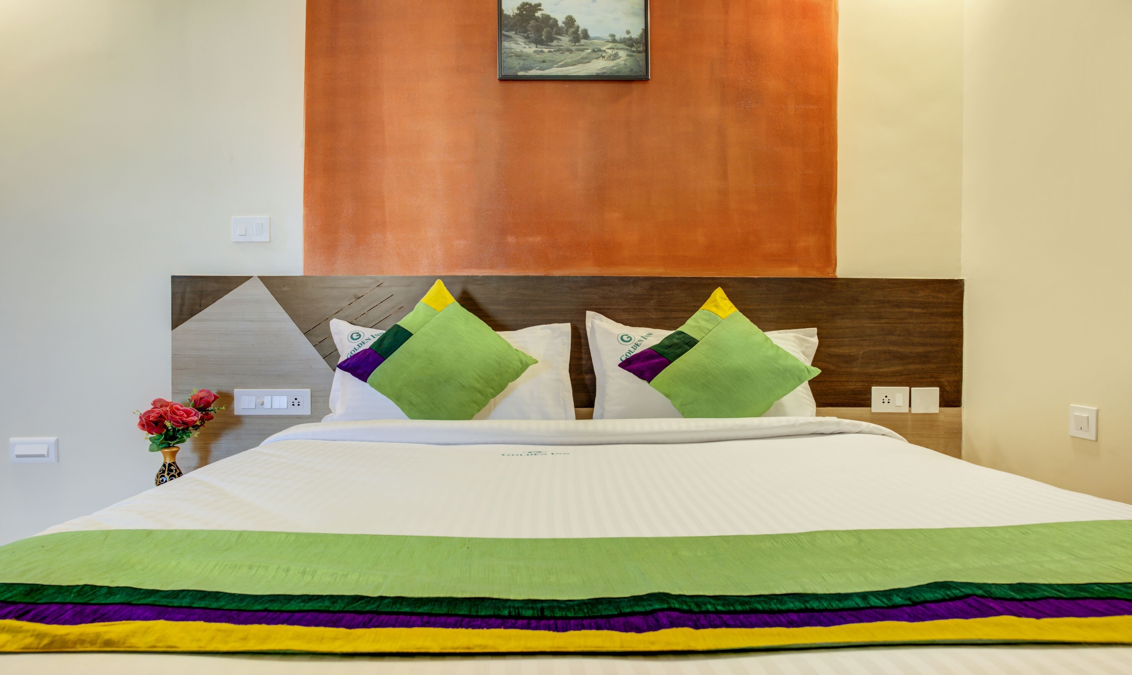 A hotel room with a neatly made bed, a wooden headboard, and a framed picture above the bed.