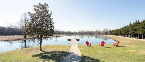 A serene lakeside area with a dock, a walking path, and three red Adirondack chairs.