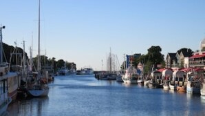 Een jachthaven met boten afgemeerd langs het water, gebouwen met rode daken, en een heldere lucht.
