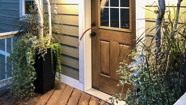 A wooden front door with a glass panel, a porch with wooden decking, and a black planter with green plants. - Beautiful lake house with pontoon INCLUDED! (Vergas)