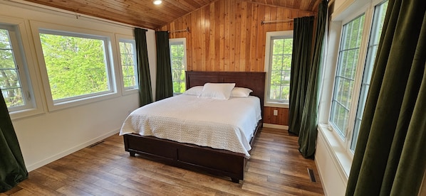 A bedroom with a wooden ceiling, a large bed, and multiple windows with curtains.
