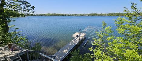 A wooden dock extending into a calm lake with clear water, surrounded by trees and a distant shoreline.