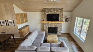 A modern living room with a stone fireplace, a large grey sectional sofa, a wooden dining table, and a large window with a view of trees. - Beautiful lake house with pontoon INCLUDED! (Vergas)