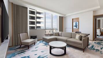 Una habitación de hotel moderna con un ventanal grande, un sofá, una silla, una mesita de centro y un televisor.