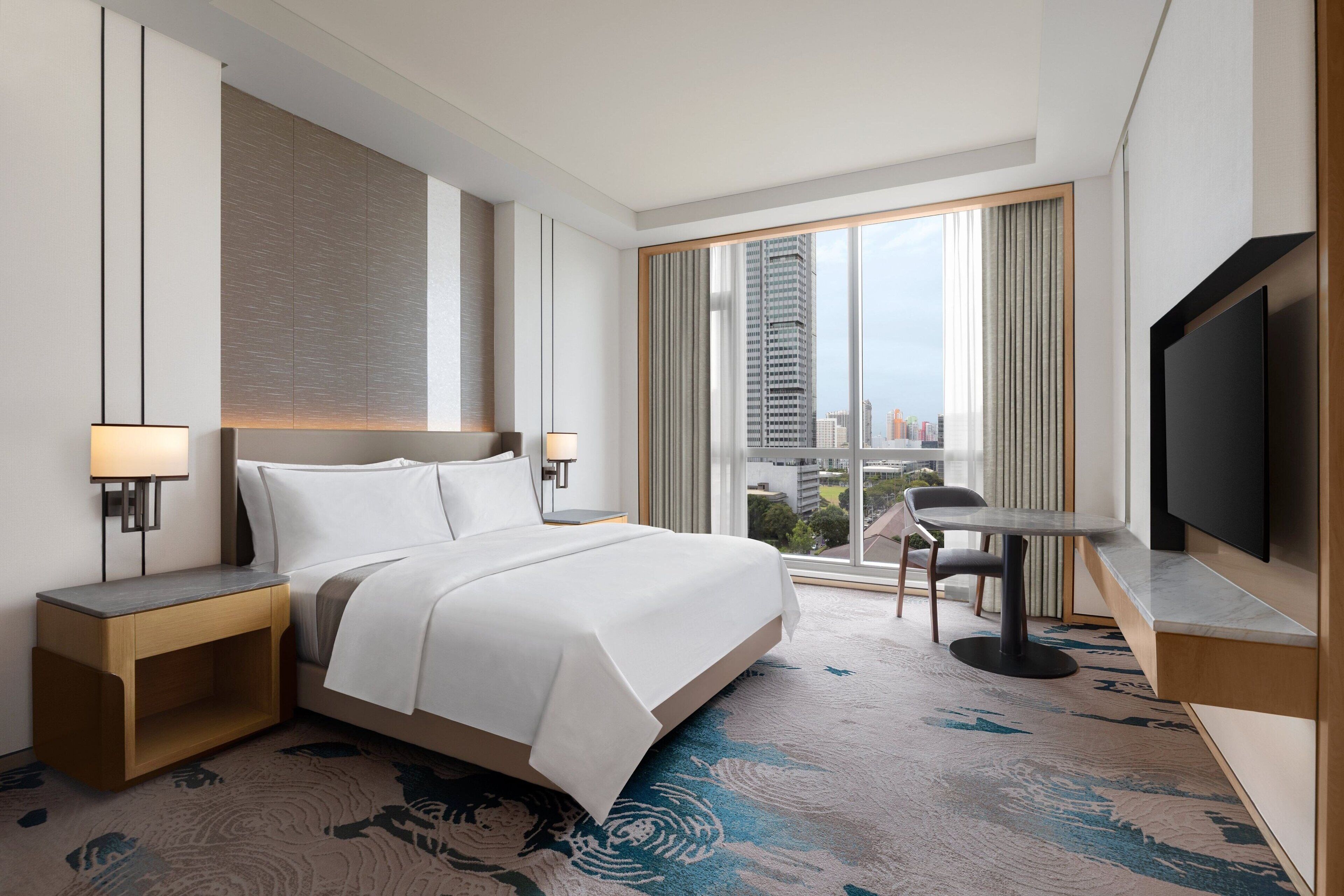 Una habitación de hotel moderna con una cama grande, un televisor de pantalla plana, una mesita pequeña y vistas a la ciudad a través de grandes ventanales.