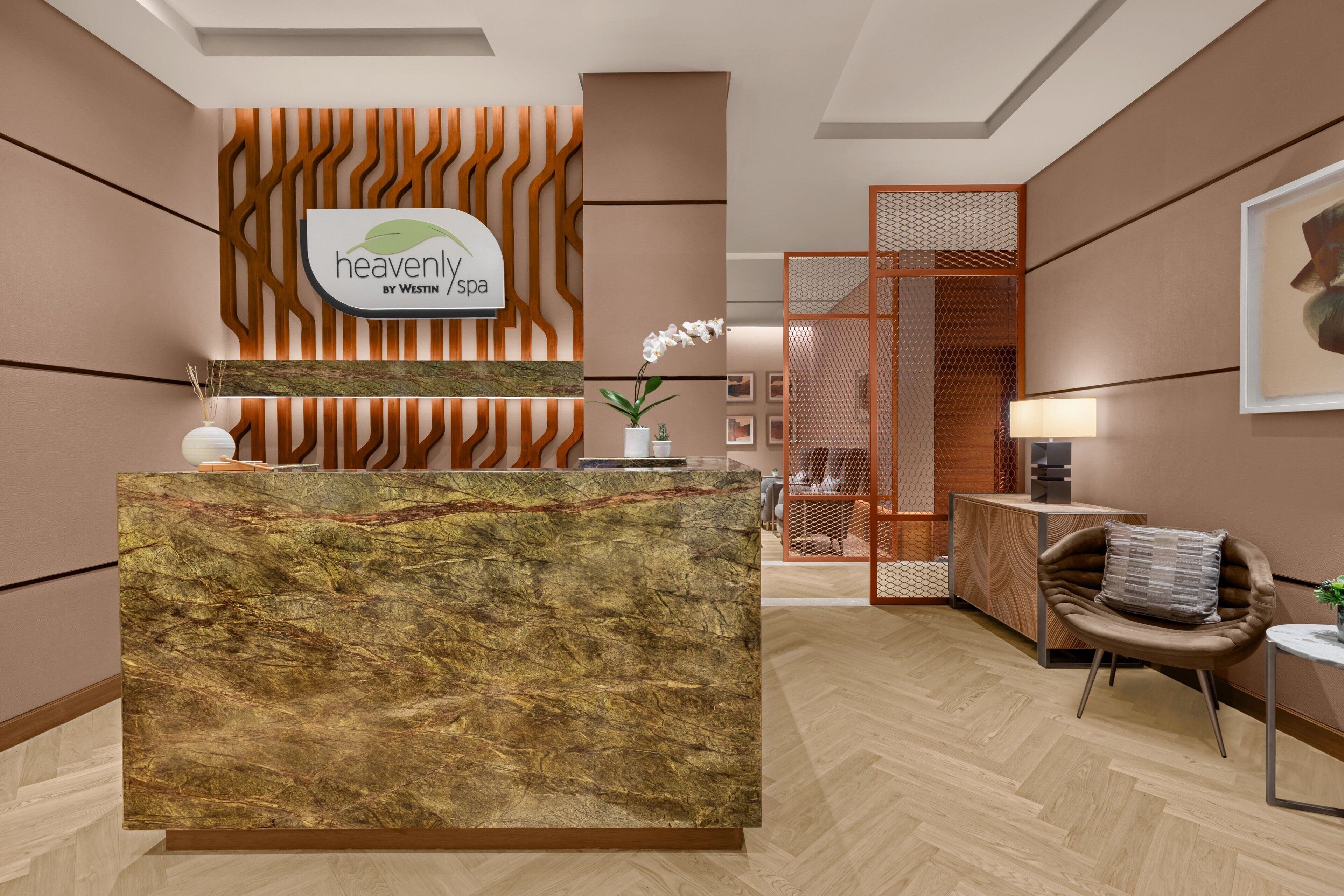 Zona de recepción con un mostrador de mármol, un cartel que dice "heavenly spa by Westin" y una decoración moderna con paneles de madera y un área de descanso con una silla y una mesita.