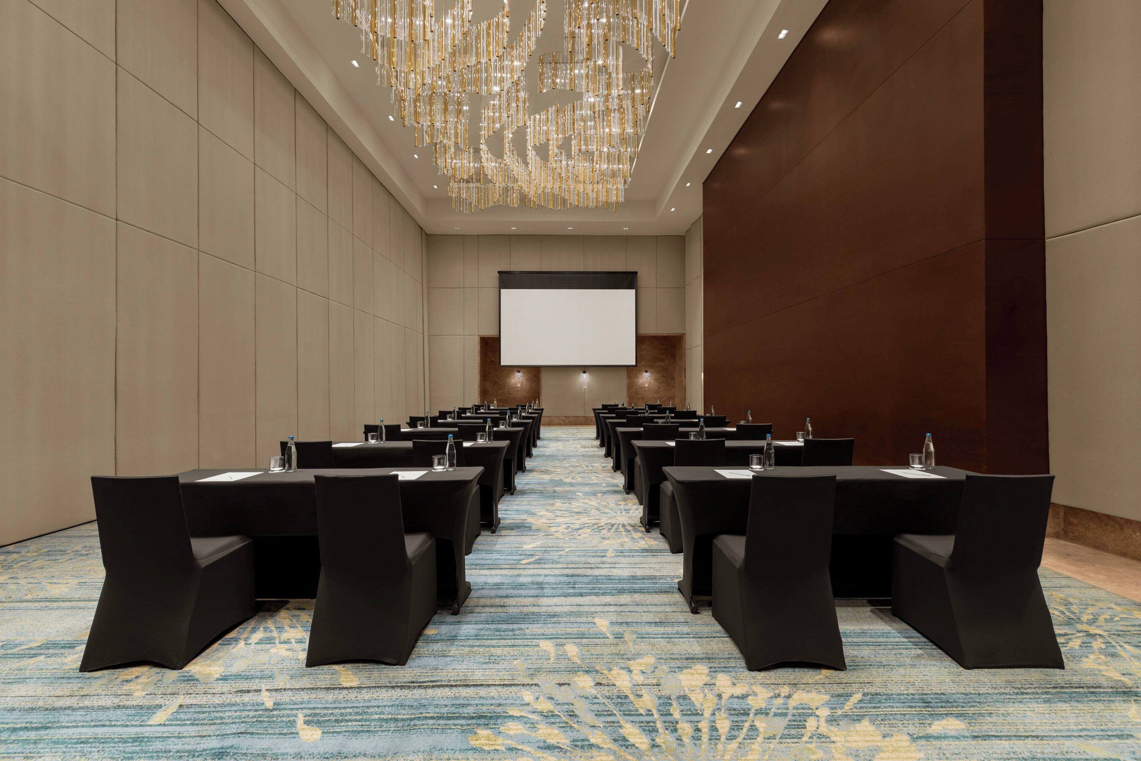 Un salón de conferencias con filas de mesas y sillas, una pantalla grande al frente y un candelabro sobre la cabeza.