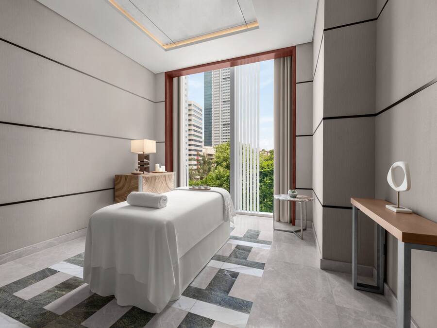 A modern hotel room with a large bed, a desk, a chair, a lamp, and a view of the city.