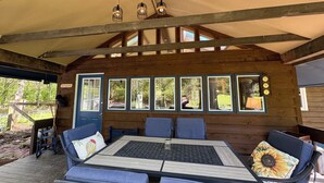 A wooden cabin with a dining area featuring a table and blue chairs, large windows, and a hanging light fixture.