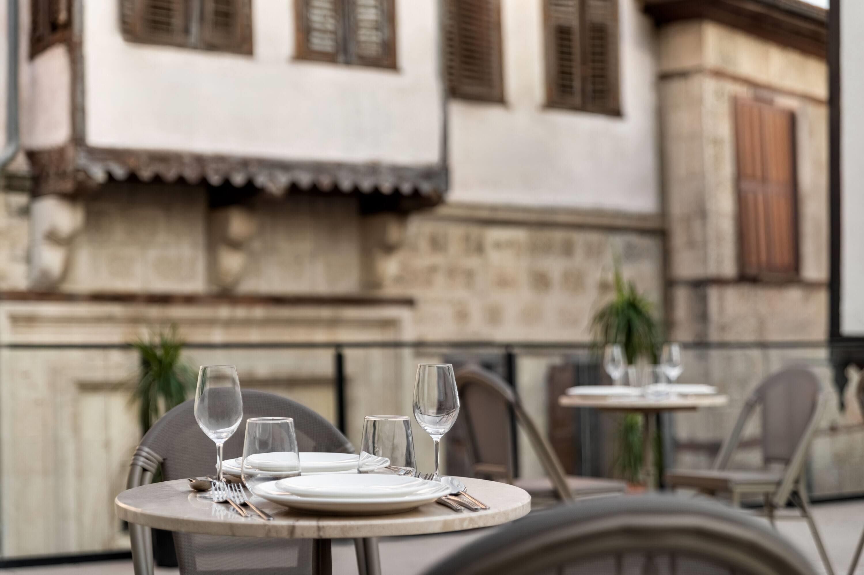 Mesa preparada para comer con platos, cubiertos y copas de vino, ubicada frente a un edificio de paredes de piedra y contraventanas de madera.