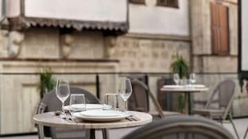 Mesa preparada para comer con platos, cubiertos y copas de vino, ubicada frente a un edificio de paredes de piedra y contraventanas de madera.