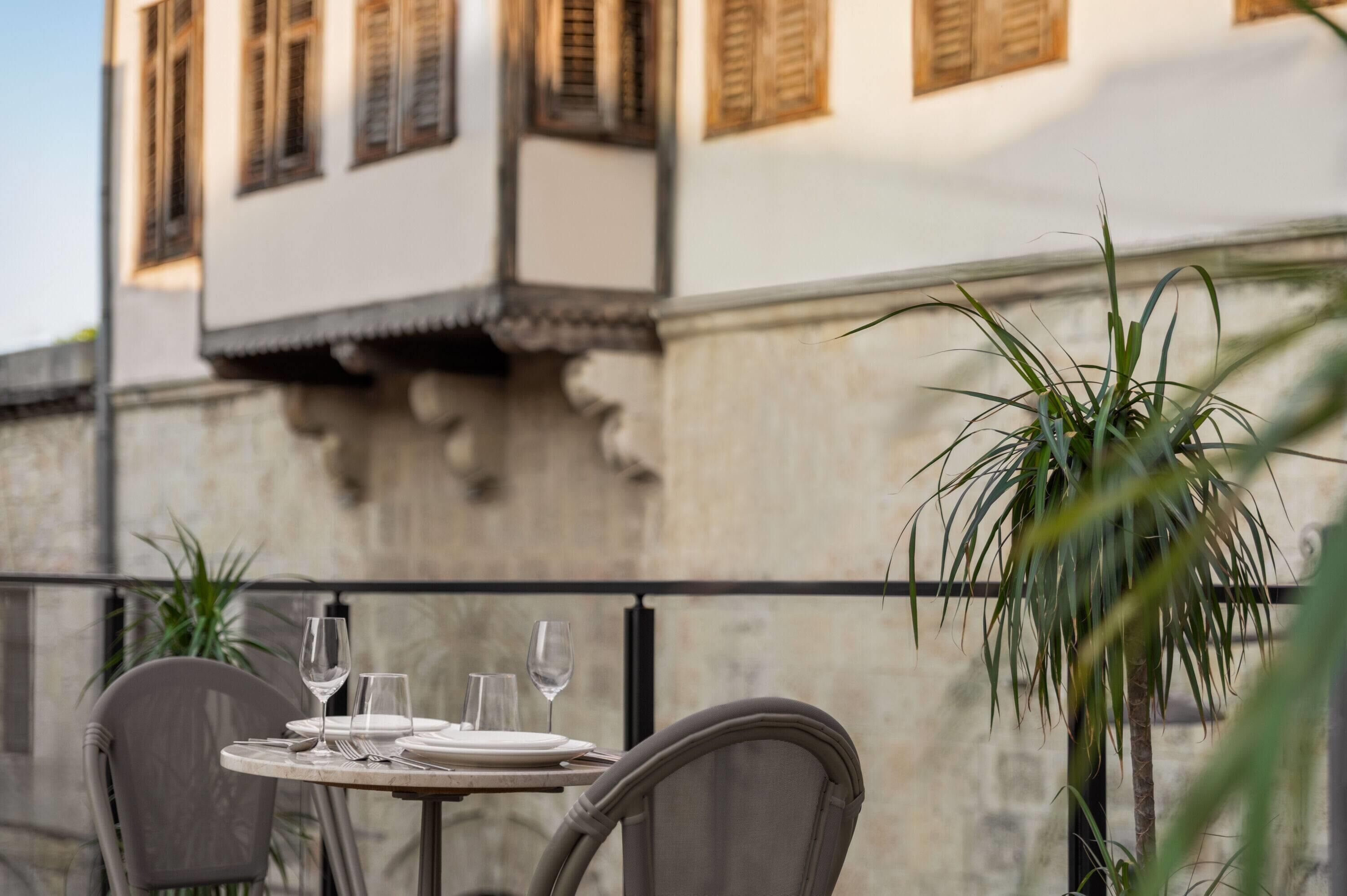 A table set for two with wine glasses and plates, a chair, and a potted plant.