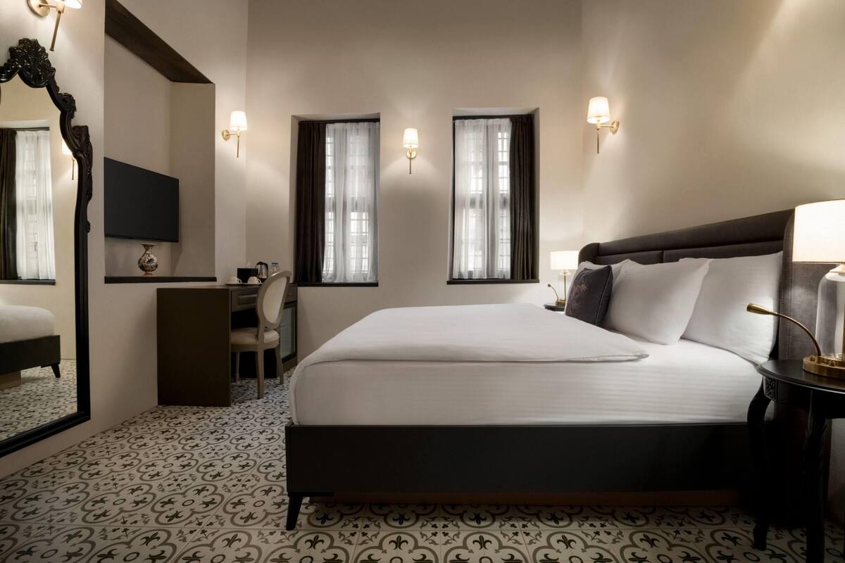 A hotel room with a large bed, a desk, a chair, a TV, and two windows with curtains.