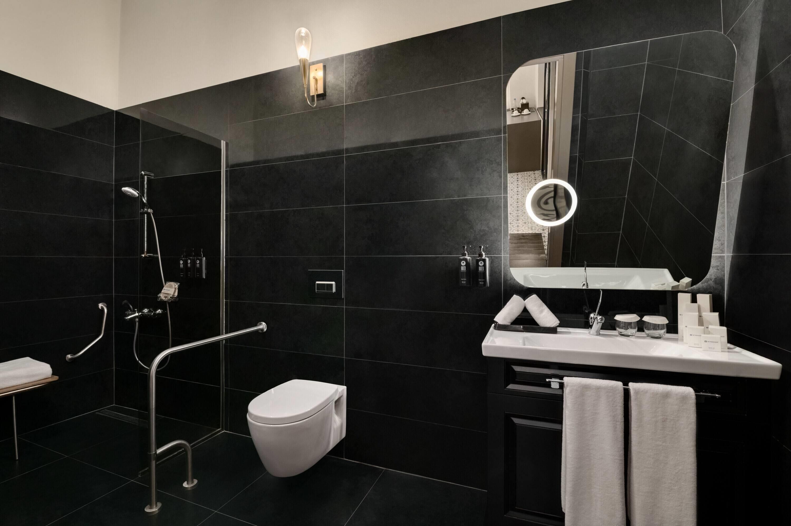 A modern bathroom with a black tiled wall, a white toilet, a sink with a mirror, and a shower area with a handheld showerhead.