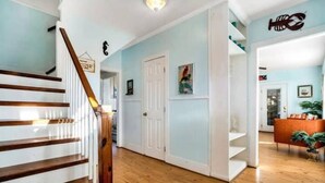 Una escalera con peldaños de madera y paredes blancas, que lleva a una habitación con una puerta blanca y un estante con un cuadro enmarcado.