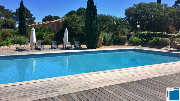 Et rektangulært swimmingpool med en træterrasse, omgivet af grønt og et par liggestole med parasoller.
