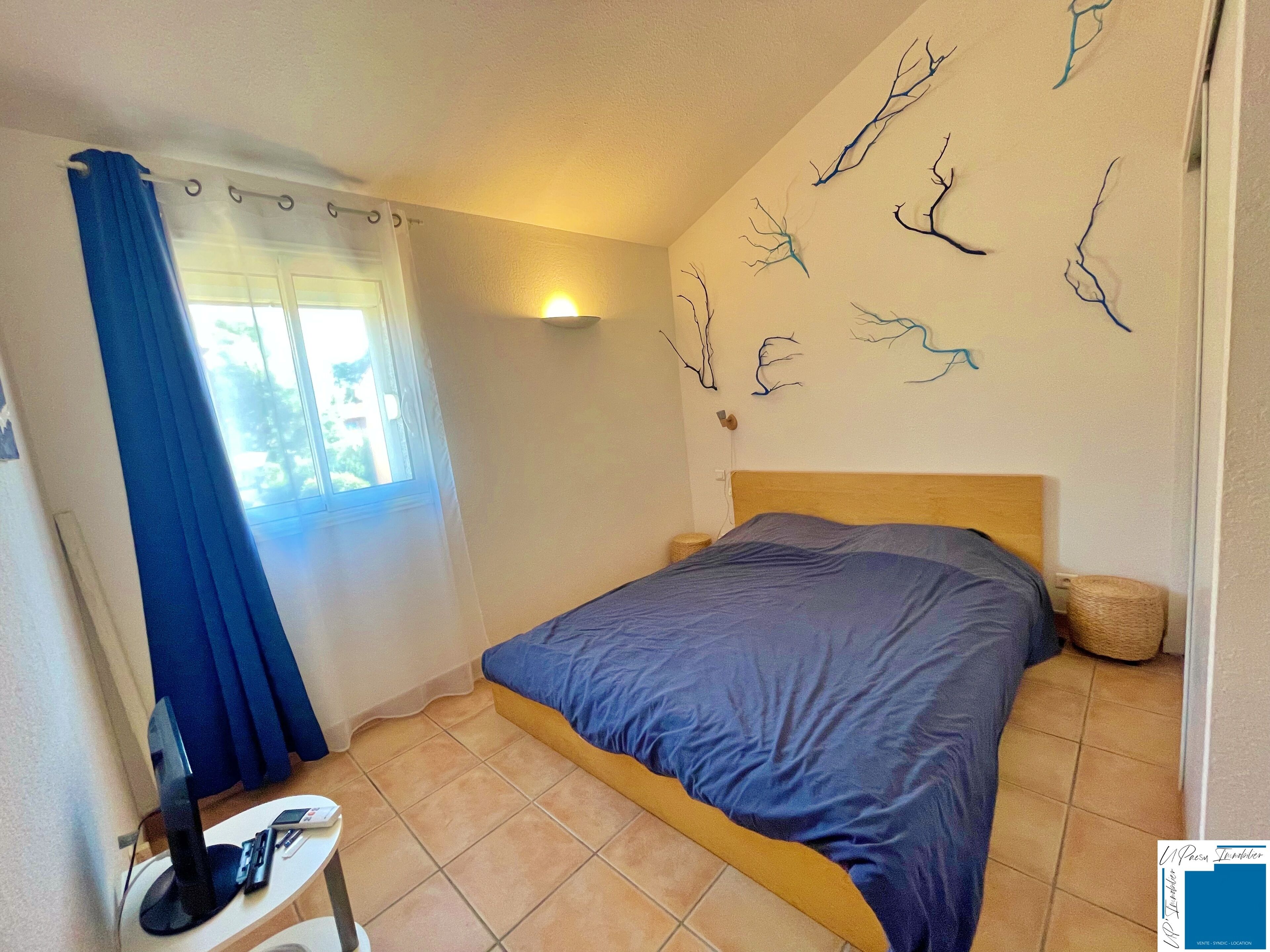 Ein Schlafzimmer mit einem Bett, einem Nachttisch, einer Lampe, einem Fenster mit Vorhängen und Wandbildern.